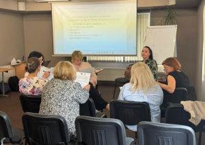 Mācību dalībnieki sēž pie galdiem, sadalīti mazākās grupās, un veic grupu darbu, piezīmējot informāciju. Priekšā redzams prezentācijas ekrāns ar tekstu