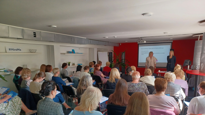 Daudz cilvēku sēž zālē un skatās uz priekšā esošo ekrānu, kurā tiek projicēta prezentācija. Priekšplānā pie ekrāna stāv divi pasniedzēji