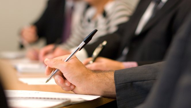 Attēlā attēlota sanāksmju telpa ar galdu, pie kura atrodas cilvēks, kurš raksta ar pildspalvu