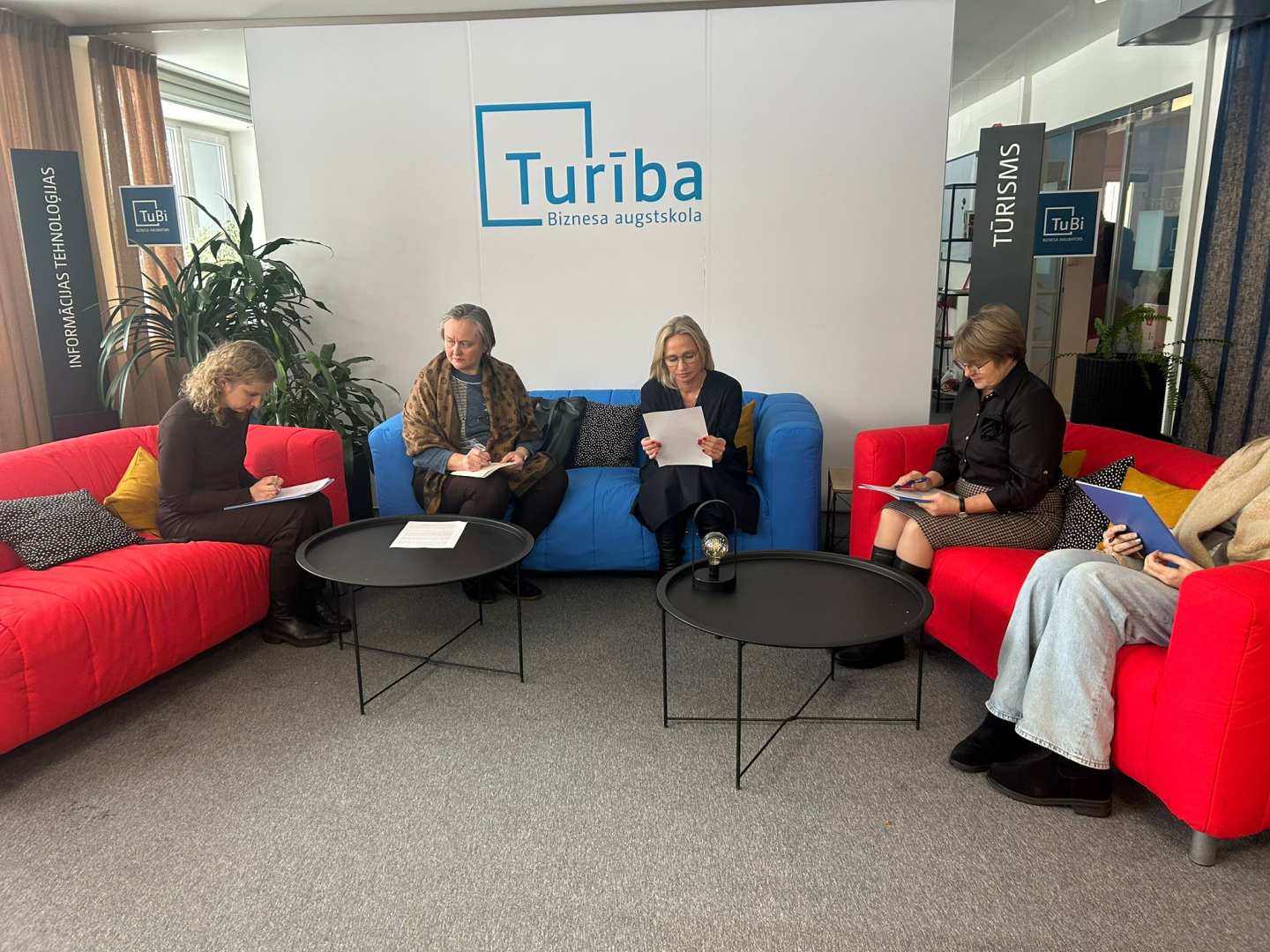 Attēlā redzami dalībnieki, kas seiž sarkanā dīvāniņā un pilda mācību uzdevumu