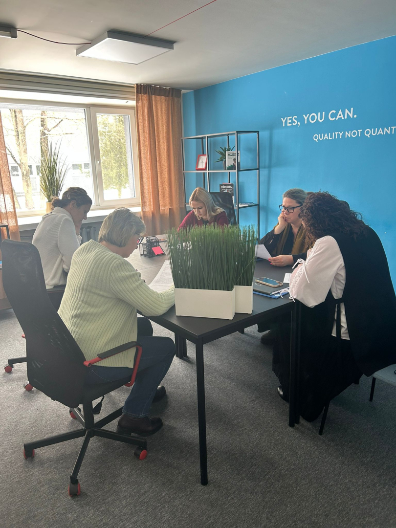 Attēlā redzami dalībnieki, kas seiž pie galdiem un pilda mācību uzdevumu