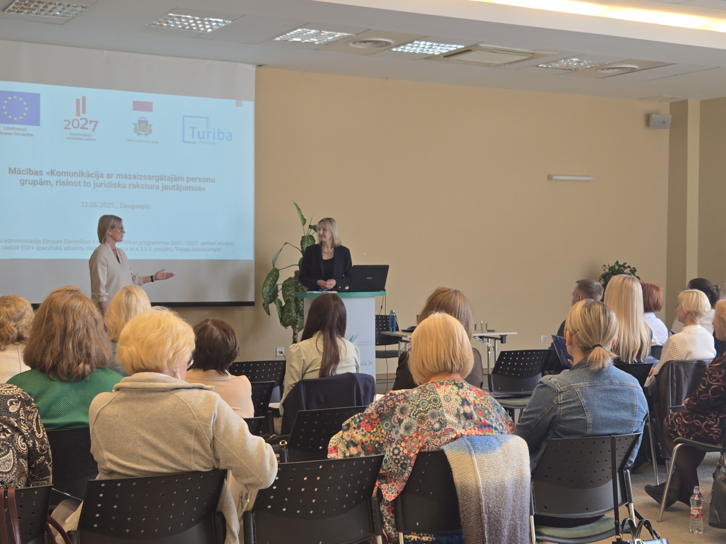 Telpa ar dalībniekiem, kas sēž rindās. Priekšplānā pie ekrāna, kurā redzams uzraksts 'Mācības: Komunikācija ar mazaizsargātajiem', stāv divas pasniedzējas