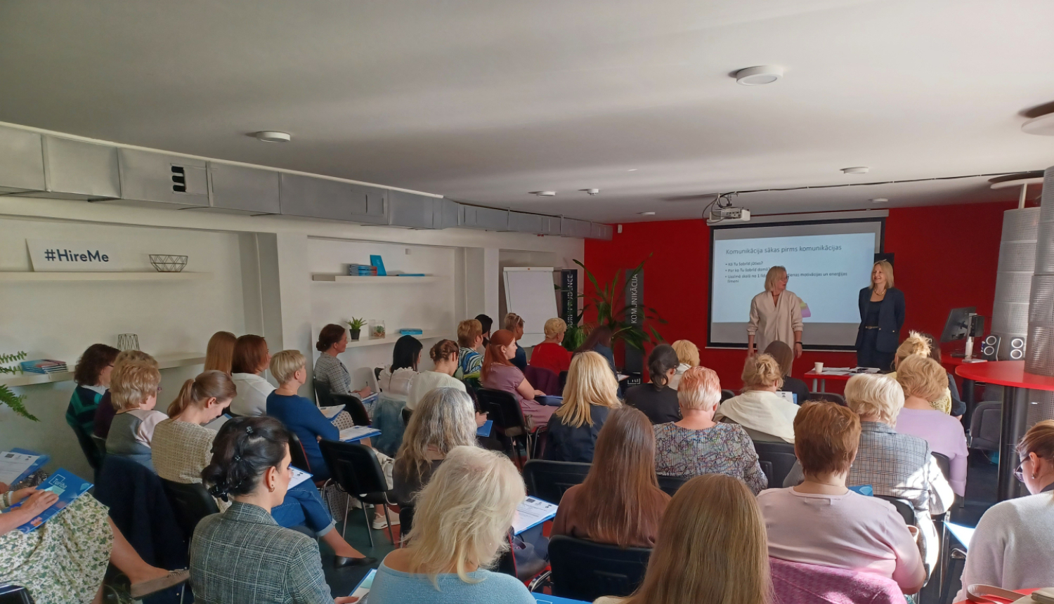 Daudz cilvēku sēž zālē un skatās uz priekšā esošo ekrānu, kurā tiek projicēta prezentācija. Priekšplānā pie ekrāna stāv divi pasniedzēji