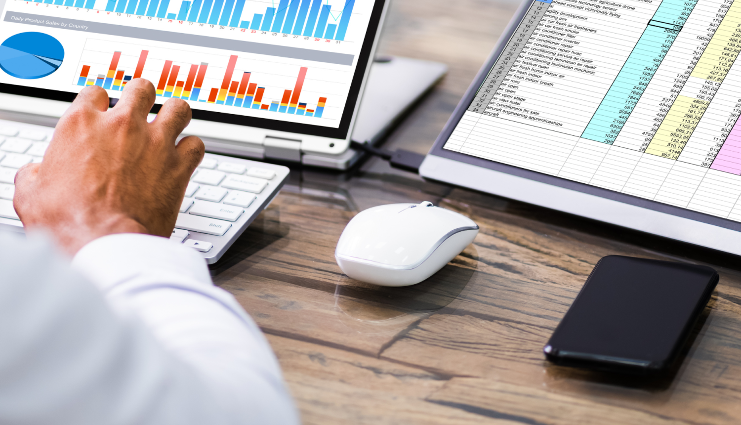 Attēlā atspoguļots galds, pie kura seiž vīrietis, kurš datorā veido grafikus