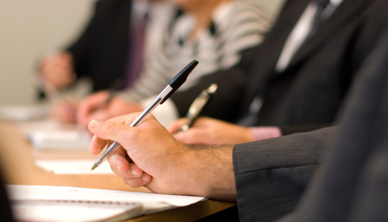 Attēlā attēlota sanāksmju telpa ar galdu, pie kura atrodas cilvēks, kurš raksta ar pildspalvu